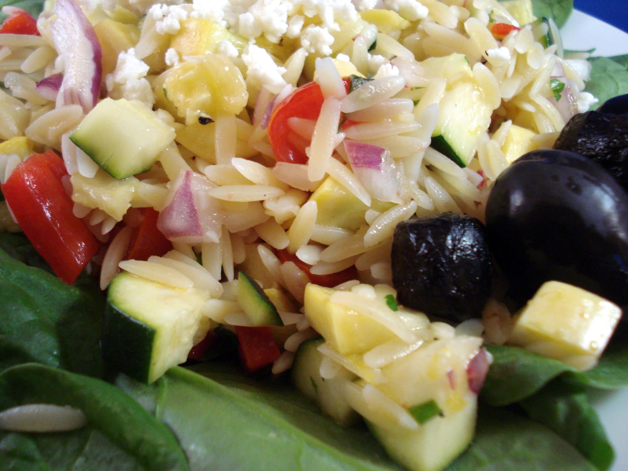 Close up of orzo on top of baby spinach, zucchini, squash and olives