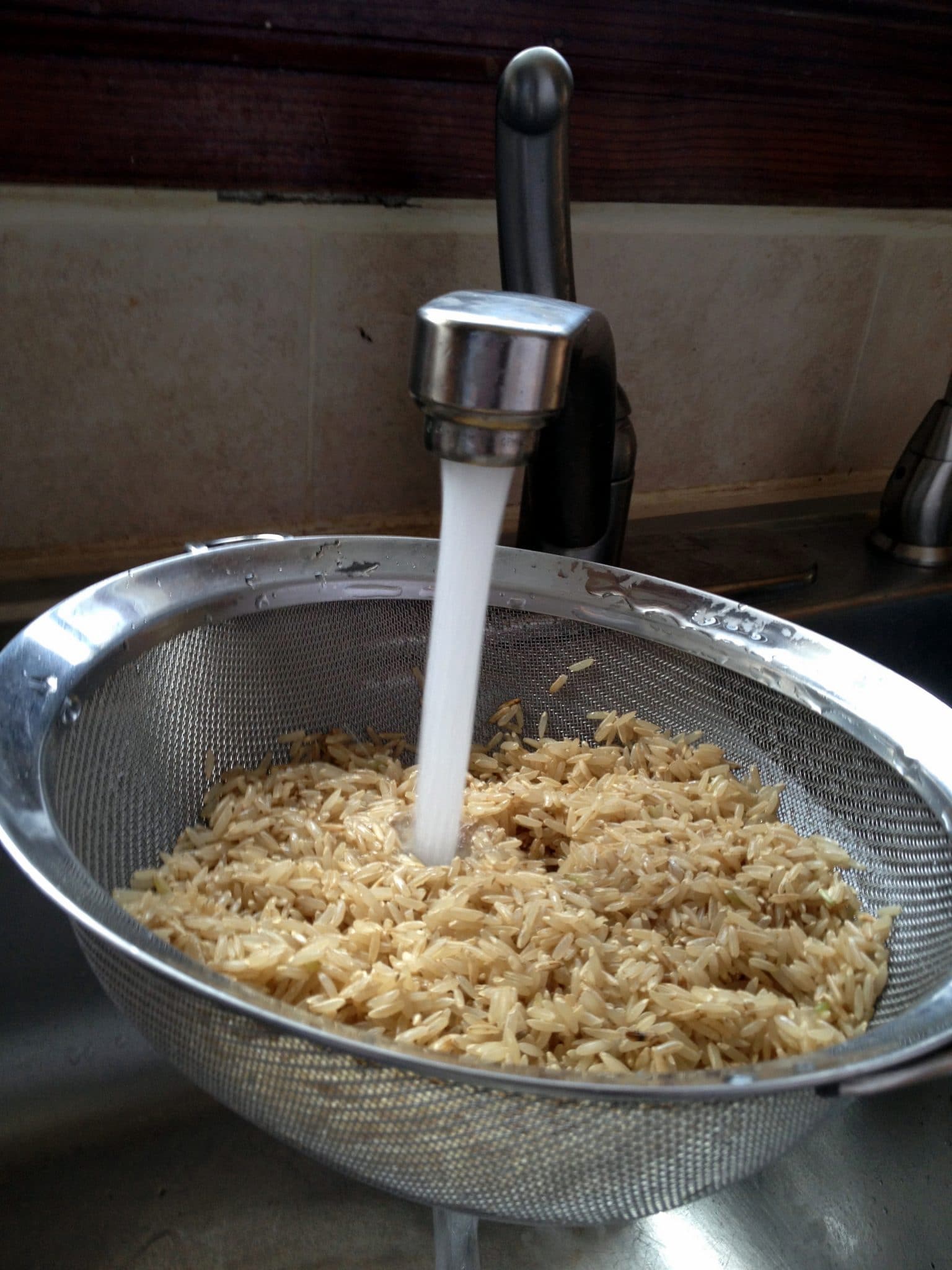 Rice in mesh strainer being rinsed by water streaming from faucet