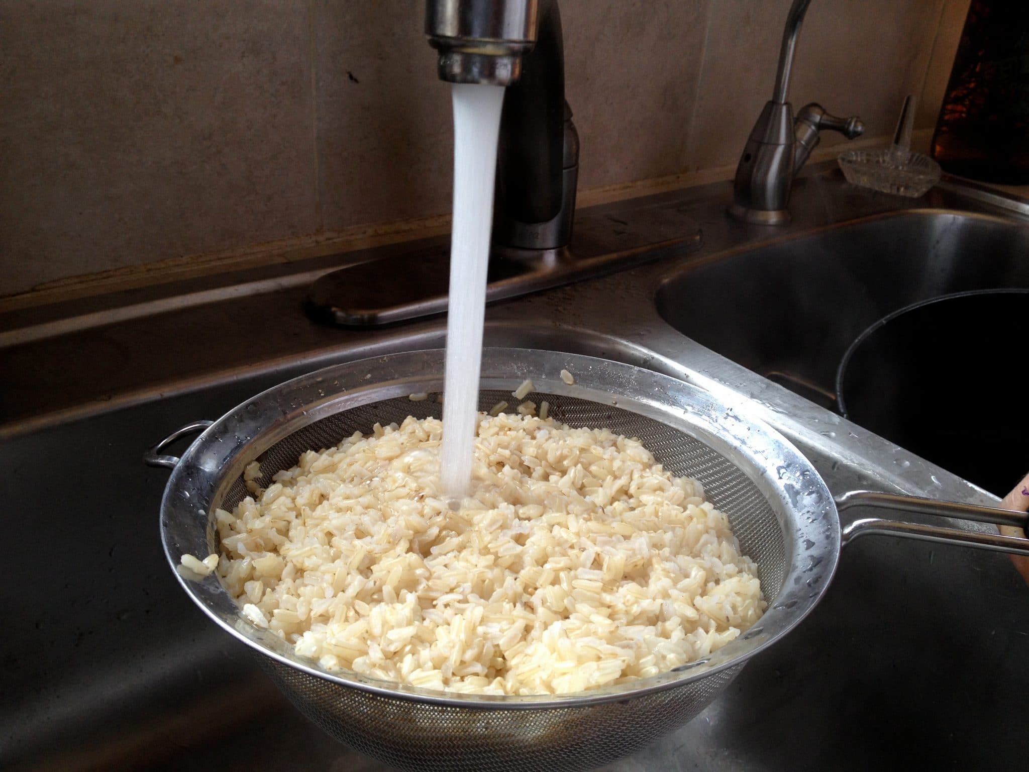 Cooked rice in mesh strainer being rinsed under hot water in sink